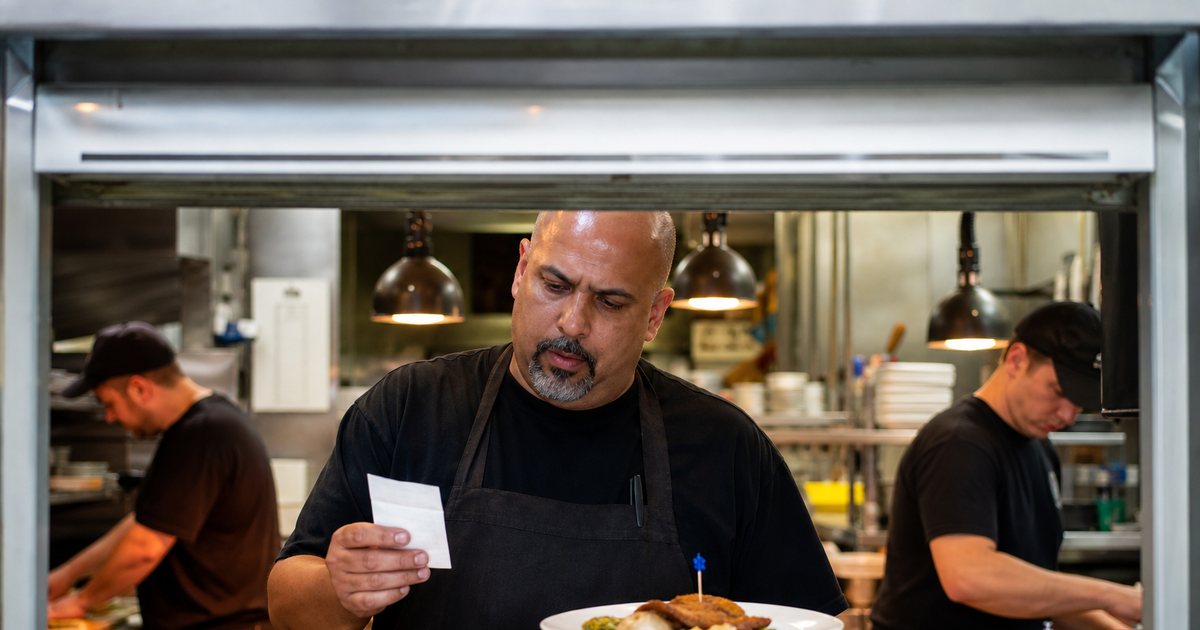 Expediter in a black apron reading a printed order ticket at a stainless steel kitchen pass window, a plated dish with a small colored pick in front, line cooks working in the background under warm heat lamps