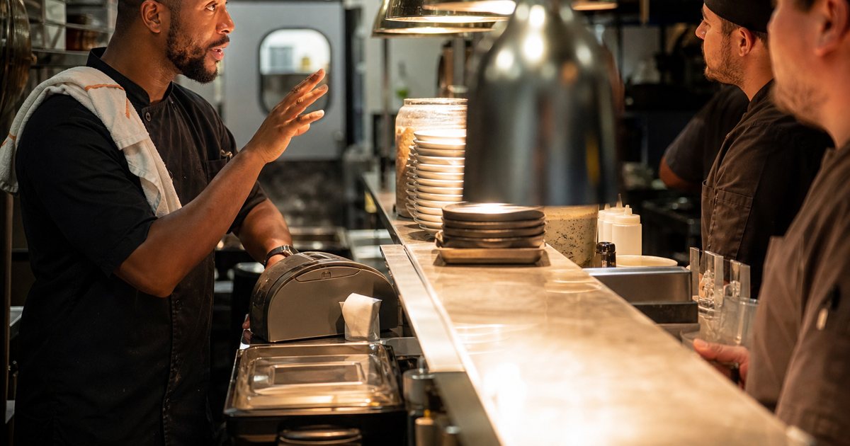 Expediter with a towel over one shoulder calling out across a stainless steel pass to a row of line cooks, one hand raised mid-gesture, ticket printer beside them, warm heat lamps overhead in a busy kitchen