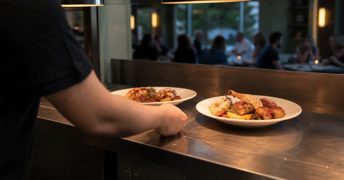 Two finished plates of roasted chicken and vegetables sitting under warm heat lamps on a stainless steel pass, a food runner in a black shirt reaching to pick them up, dining room blurred beyond the window