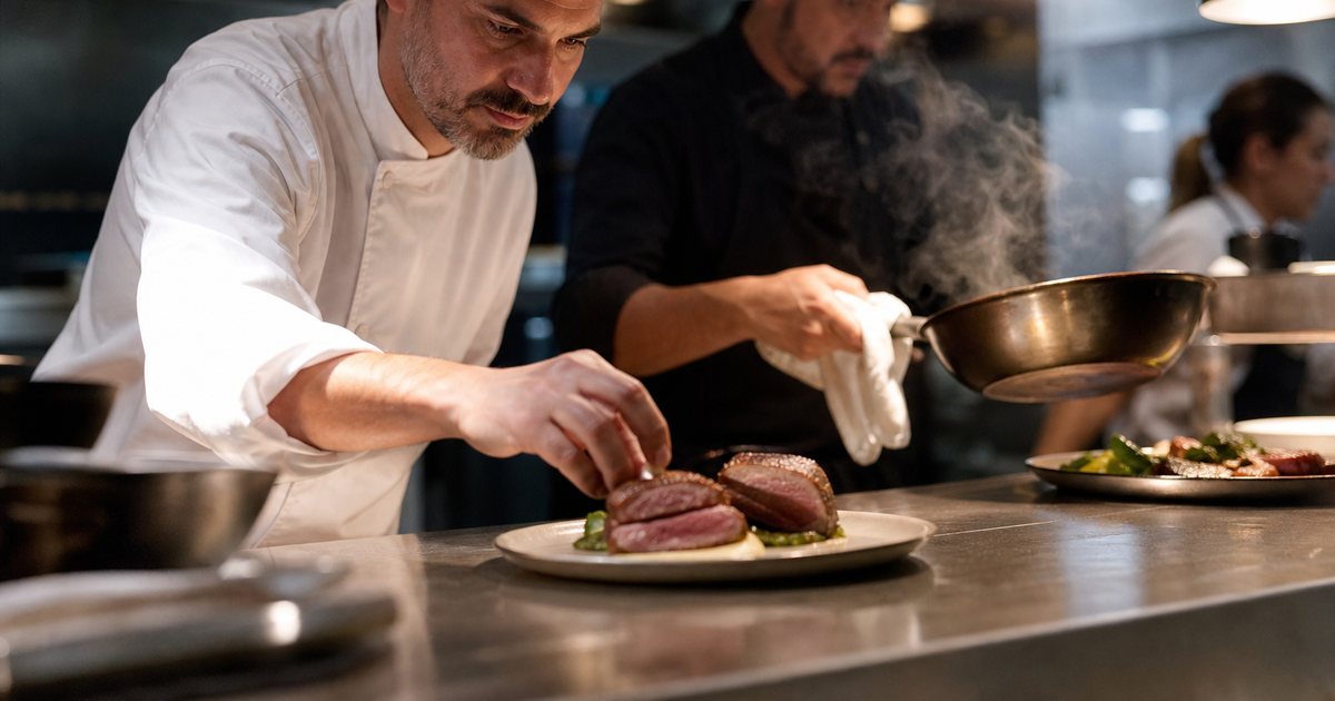 Chef in a white coat plating a seared steak at a stainless steel pass under warm heat lamps while a line cook reaches in with a saute pan, steam rising, shallow depth of field in a busy restaurant kitchen at night
